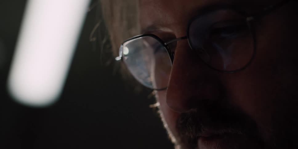 An extreme close-up shot of a focused man's glasses reflecting a digital screen as he works intently in a dark room