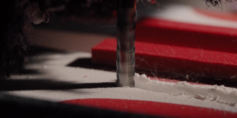 A close-up, slow-motion shot shows a CNC milling machine drill bit precisely cutting a shape into a red and white composite material, sending shavings flying