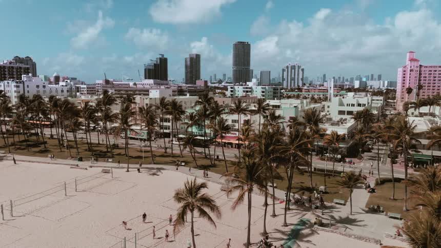 A slow aerial dolly shot moves right over Miami's South Beach, showing people playing volleyball, palm trees, and the city skyline