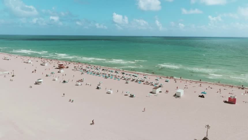 A serene, static wide shot captures the bustling activity on a beautiful beach with turquoise waters and a clear sky