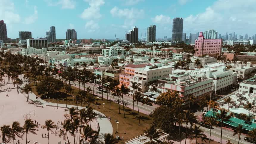 Aerial view pulls back over the sandy shores and palm trees of Miami's famous Ocean Drive, revealing the vibrant Art Deco hotels and city skyline