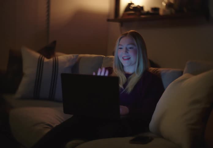 A smiling young woman waves goodbye on a video call and closes her laptop while sitting on a cozy couch at night