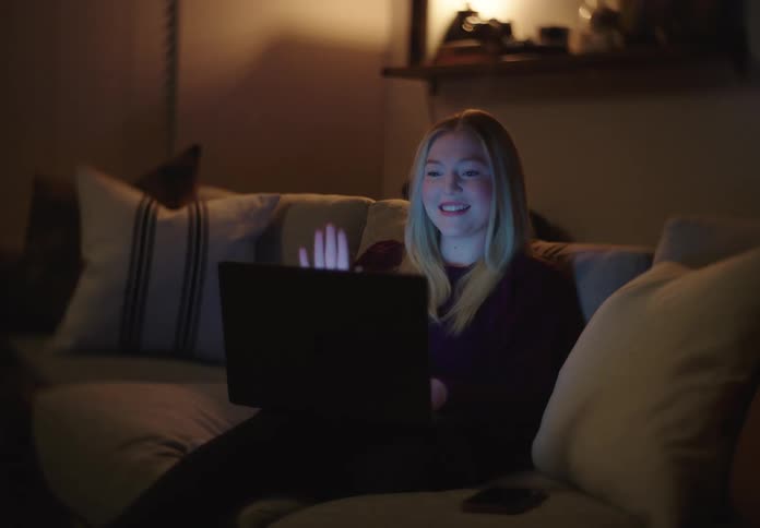 Happy young woman video chatting on her laptop at night while relaxing on the couch in a cozy living room