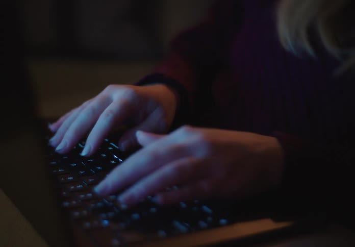 Close up of a woman's hands typing on a laptop keyboard in a dark room illuminated by the screen's glow