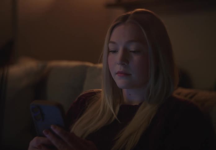 A young blonde woman relaxes on her couch at night, scrolling through her smartphone in a dark room