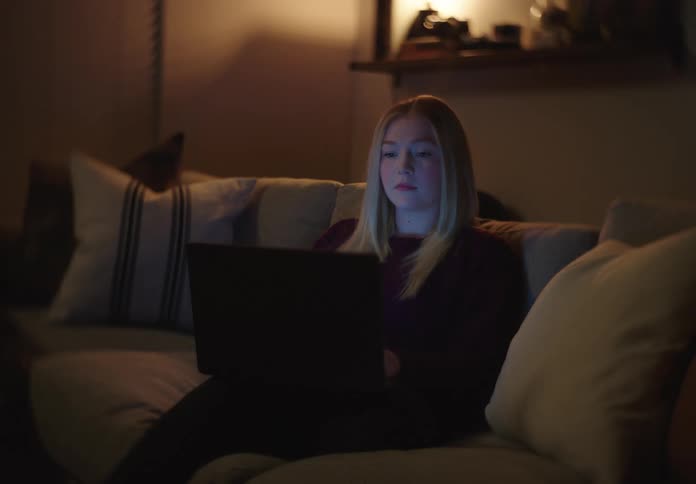 A young woman uses her laptop on the couch at night, the screen illuminating her face in the dark