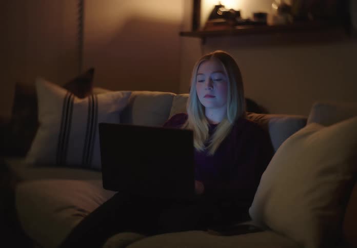 A blonde woman working on her laptop at night gets distracted by her phone while sitting on a cozy couch