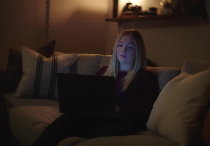 A teenage girl works on her laptop at night, her face illuminated by the glow of the screen