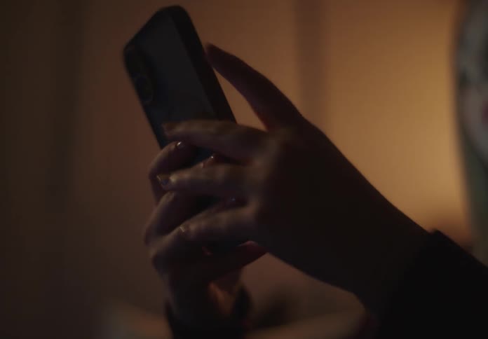 A close-up shot of silhouetted hands scrolling and texting on a smartphone in a dimly lit room at night