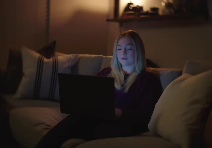 A young woman sits on a couch in a dimly lit living room at night, her face illuminated by the glow of her laptop screen as she works