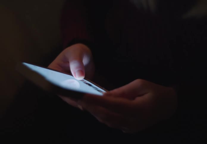 Close-up of a person's hands illuminated by their smartphone screen as they scroll and text in a dark room