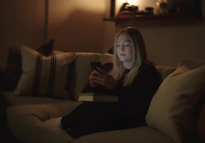 A young woman relaxes on a cozy couch at night, illuminated by the glow of her smartphone screen