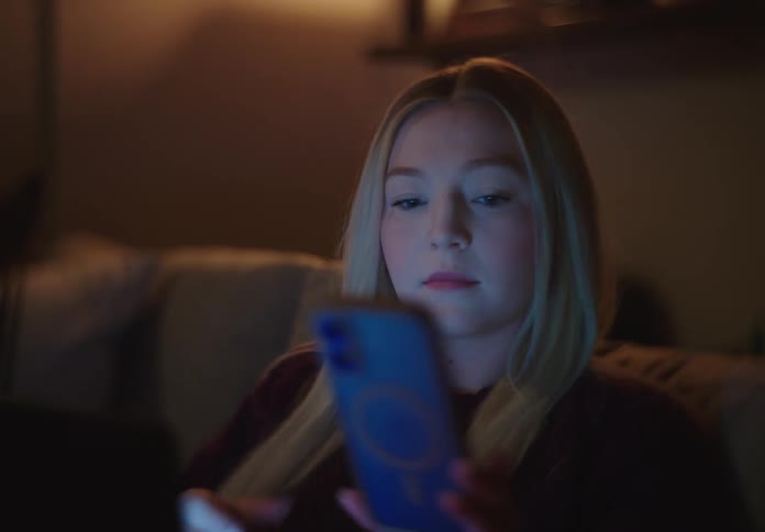 A young blonde woman works on her laptop and checks her phone while relaxing on the couch in a dimly lit room at night