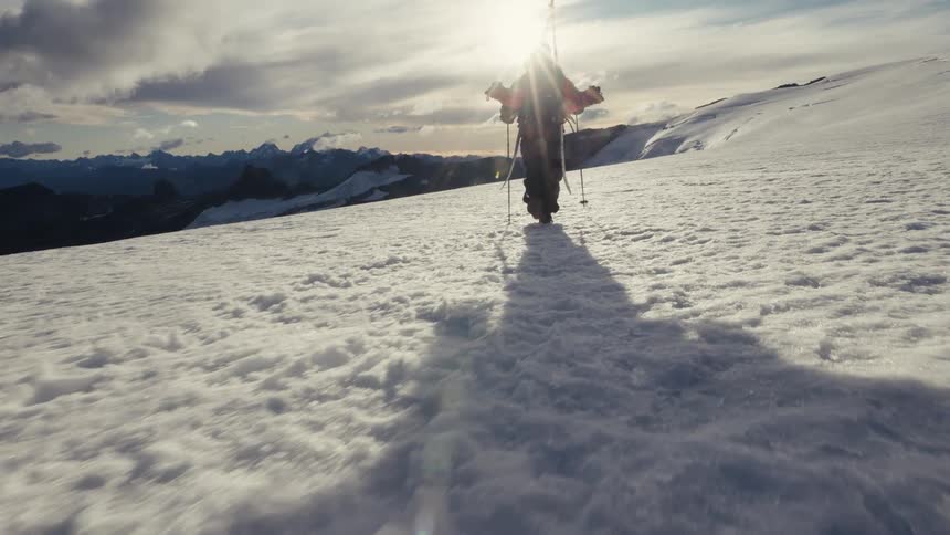 A low-angle shot follows a lone mountaineer hiking up a vast snowy slope towards the bright sun, casting a long shadow