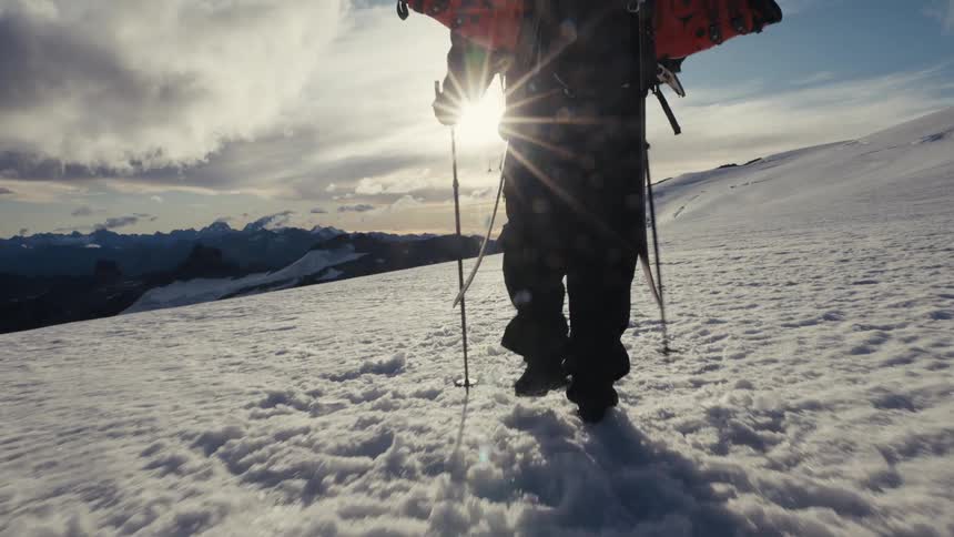 A lone skier with trekking poles hikes up a snowy mountain towards the bright morning sun