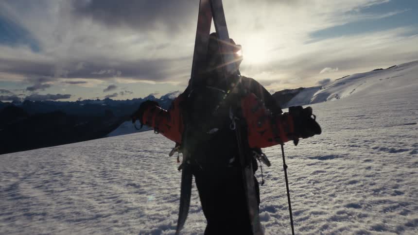 A silhouetted backcountry skier stands on a snowy mountain summit, admiring the sunset view in slow motion