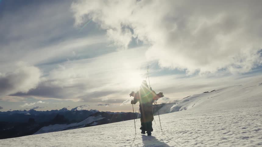 A silhouetted mountaineer carrying skis hikes up a vast snowy mountain towards the bright, brilliant sun