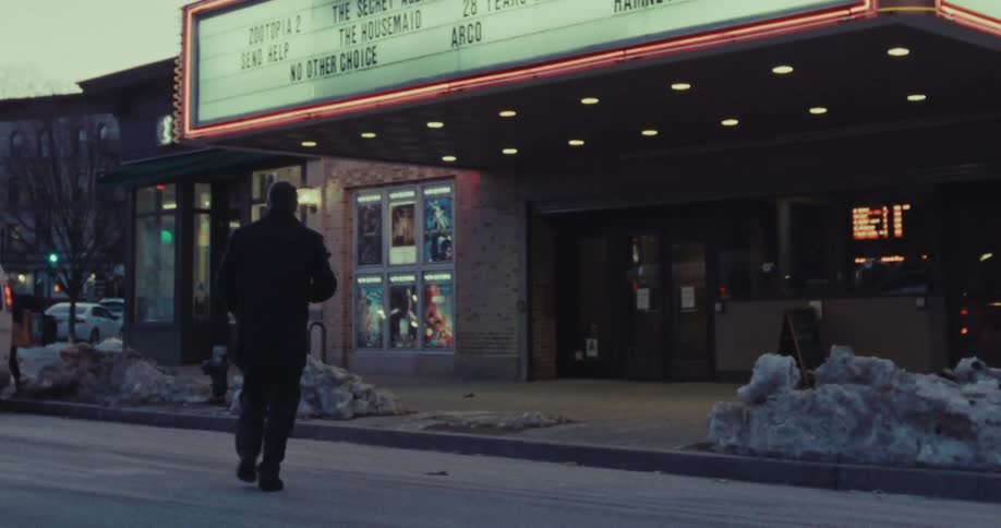 A solitary man walks down a snowy city street towards a lit movie theater marquee at dusk