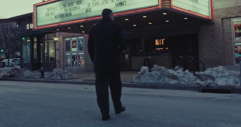 A man carrying a bouquet of flowers walks across a snowy street toward a movie theater for a date night