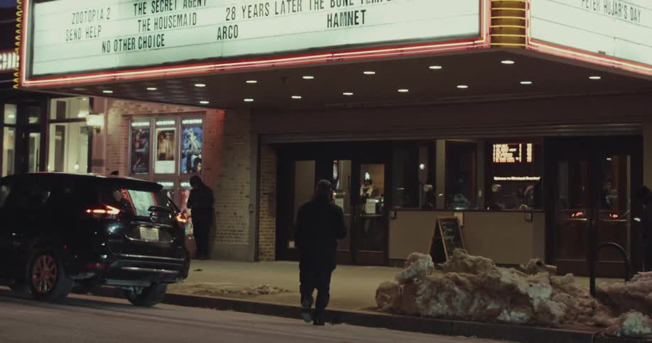 A man in a coat walks towards the brightly lit entrance of a city movie theater on a cold winter night