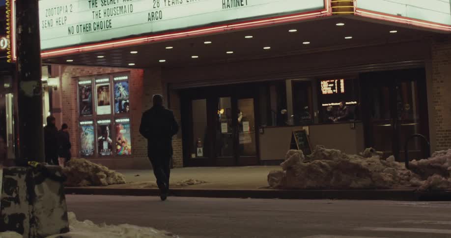 A man walks toward the entrance of a movie theater on a snowy city street at night