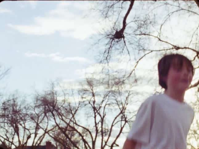 A smiling young boy in a white t-shirt happily jumps and plays outside in a park in this nostalgic, vintage-style footage
