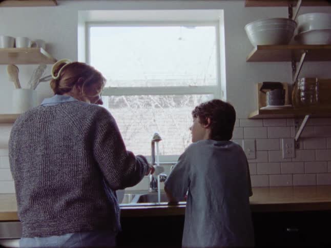 A mother and her young son stand side-by-side washing dishes together at the kitchen sink