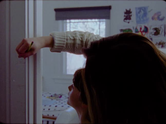 A mother lovingly marks her son's height on a door frame growth chart in a nostalgic, film-like clip