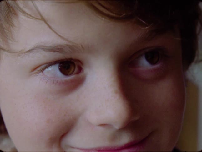 Vintage 8mm film captures a tender close-up of a young boy's freckled face as his eyes open and a gentle smile appears