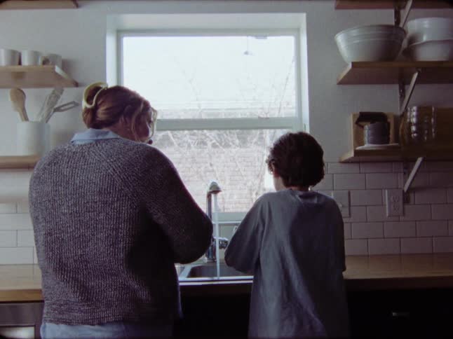 A mother and her son stand together at the kitchen sink washing dishes in their cozy home, captured in authentic film footage