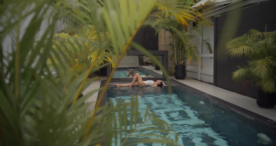 Framed by palm fronds, a woman in a white swimsuit relaxes on the edge of a modern swimming pool at a luxurious tropical villa