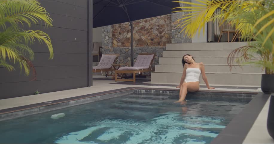 A beautiful woman in a white swimsuit soaks up the sun while sitting on the edge of a tranquil private pool at a modern tropical villa