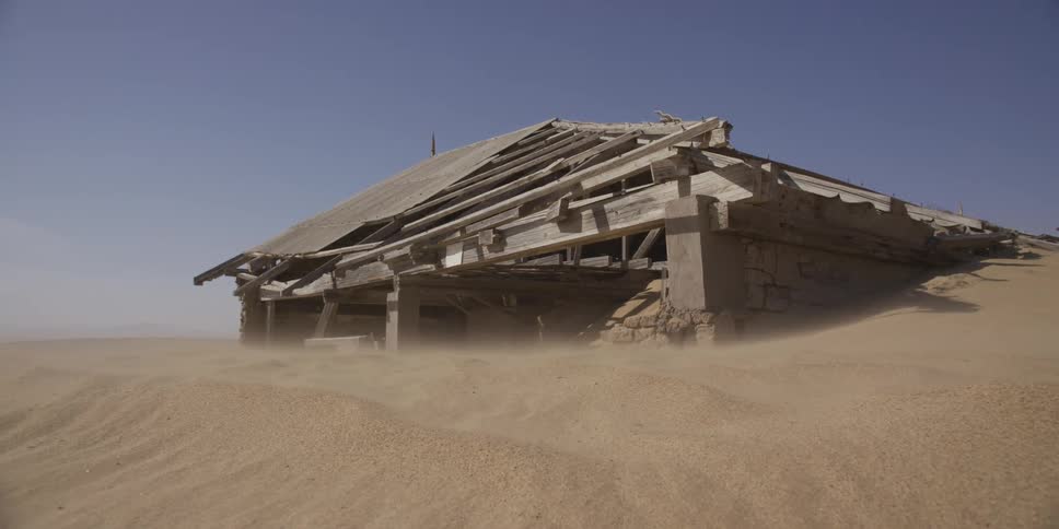 A desolate, abandoned house sinks into the desert as a strong wind blows sand across the barren landscape