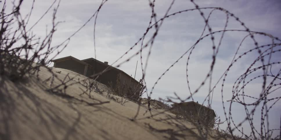 Barbed wire coils on a windswept sand dune in front of a desolate building under a hazy sky