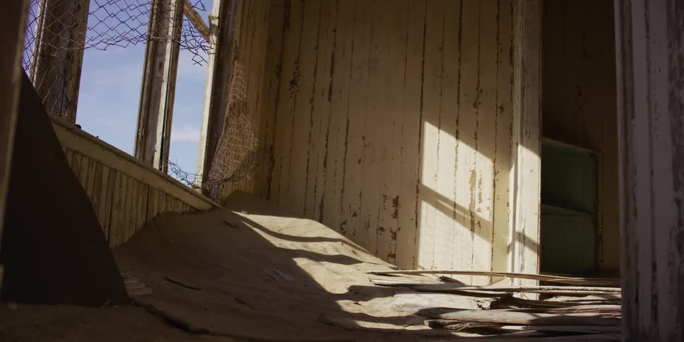 A large sand dune fills the interior of a dilapidated house in a desert ghost town, with sunlight streaming through the broken window