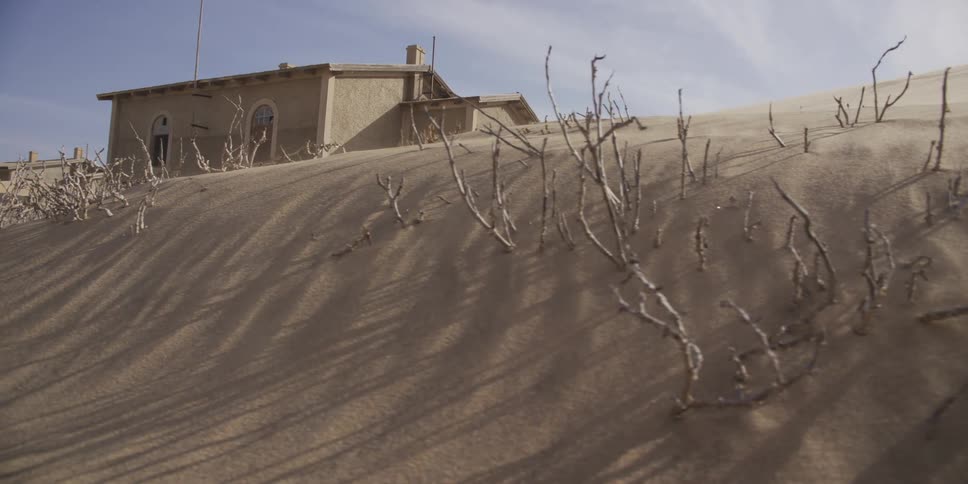 A slow tracking shot reveals an abandoned building being swallowed by a massive sand dune in a desolate desert ghost town