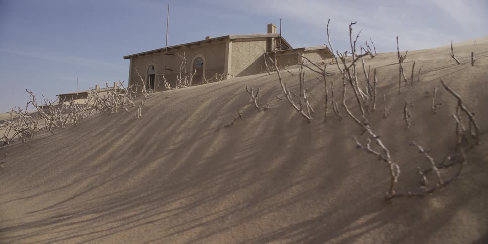 A slow pan across a sand dune reveals an abandoned building being reclaimed by the desert under a sunny sky