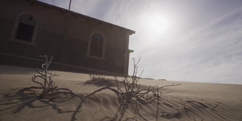 A dramatic slow-motion dolly shot captures wind blowing sand over a dune in front of an abandoned building in a desolate desert ghost town