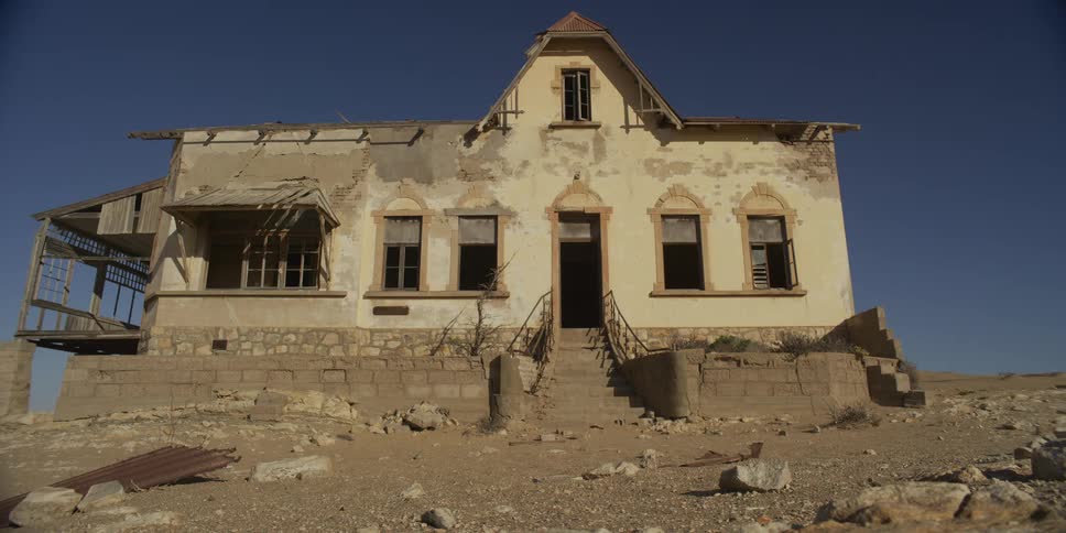 A dilapidated abandoned house sits in disrepair in a deserted wasteland under a clear blue sky