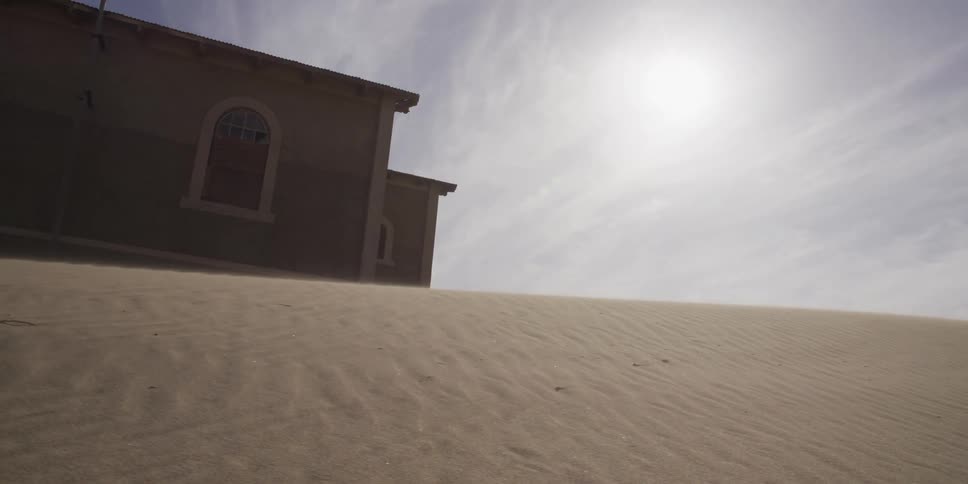 A slow dolly shot reveals an abandoned building being consumed by blowing desert sand under a harsh sun