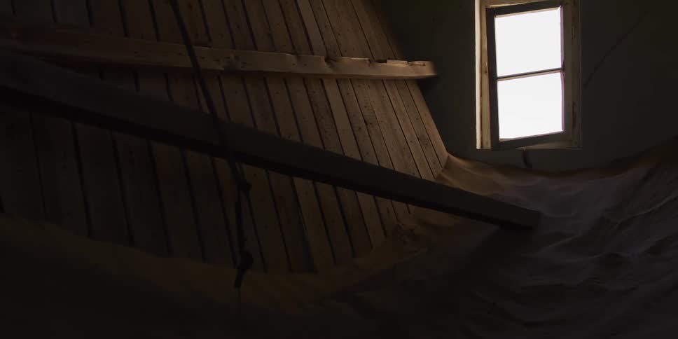 Bright light from a lone window illuminates a dark, wood-paneled room mysteriously filled with sand