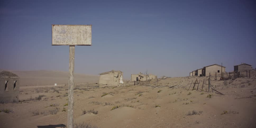 A weathered warning sign stands in a desolate desert ghost town with abandoned buildings being reclaimed by the sand