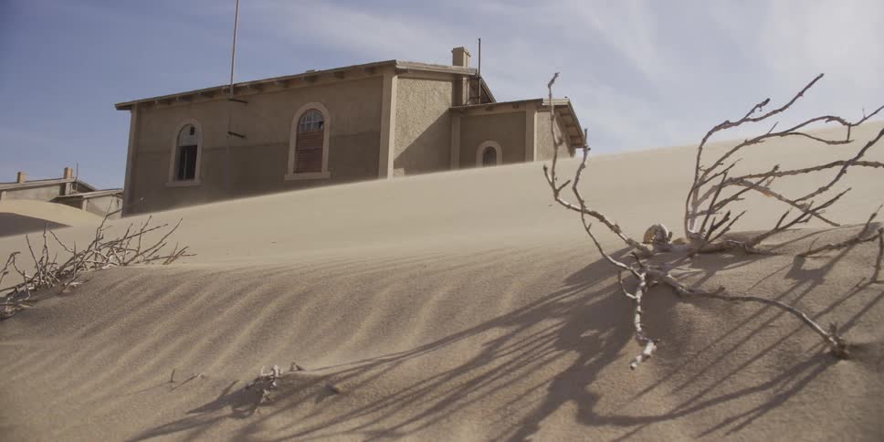 An abandoned building in a desert ghost town is slowly consumed by shifting sand dunes as the wind blows in slow motion