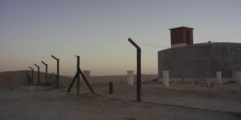 A desolate desert outpost with a dilapidated fence and a water tank stands silently at dusk