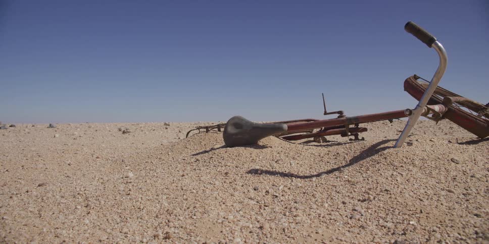 A forgotten red bicycle lies partially buried in the sand of a vast desert under a clear blue sky