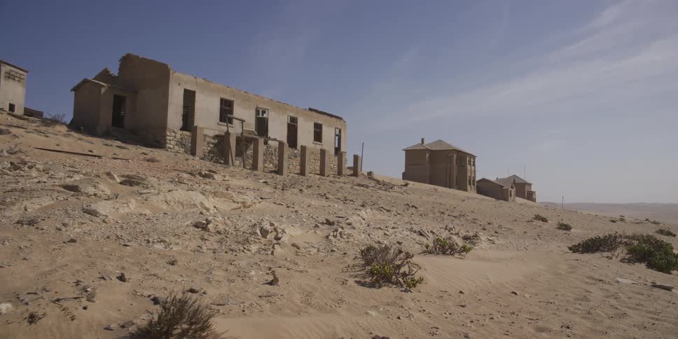 A slow tracking shot reveals the decaying ruins of a ghost town being reclaimed by the desert sand