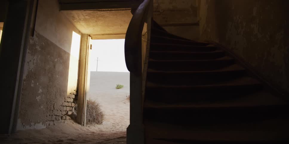 Slow pull-back shot revealing a dark, sand-filled staircase inside an abandoned desert home in Kolmanskop, Namibia