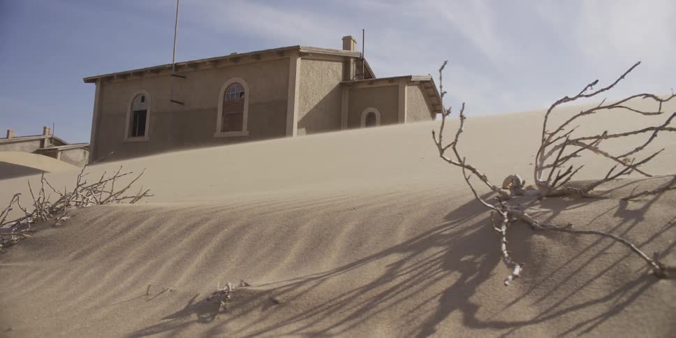 A slider shot reveals a dead bush on a windy sand dune in front of an abandoned building in a desert ghost town