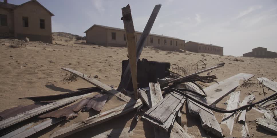 Weathered wooden planks and debris are strewn across the sand in a desolate, abandoned desert town