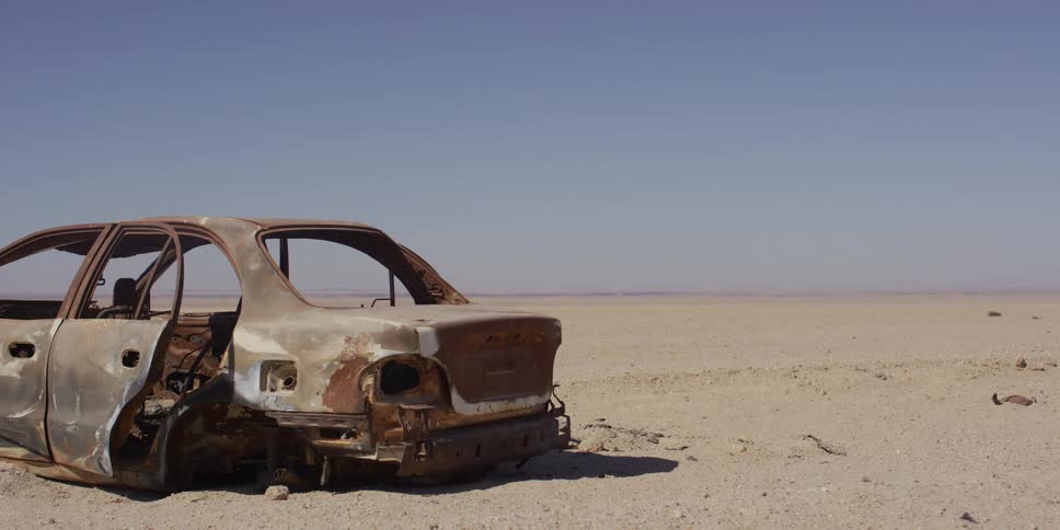 The rusted and burnt-out shell of an abandoned car sits motionless in a vast, empty desert on a hot sunny day